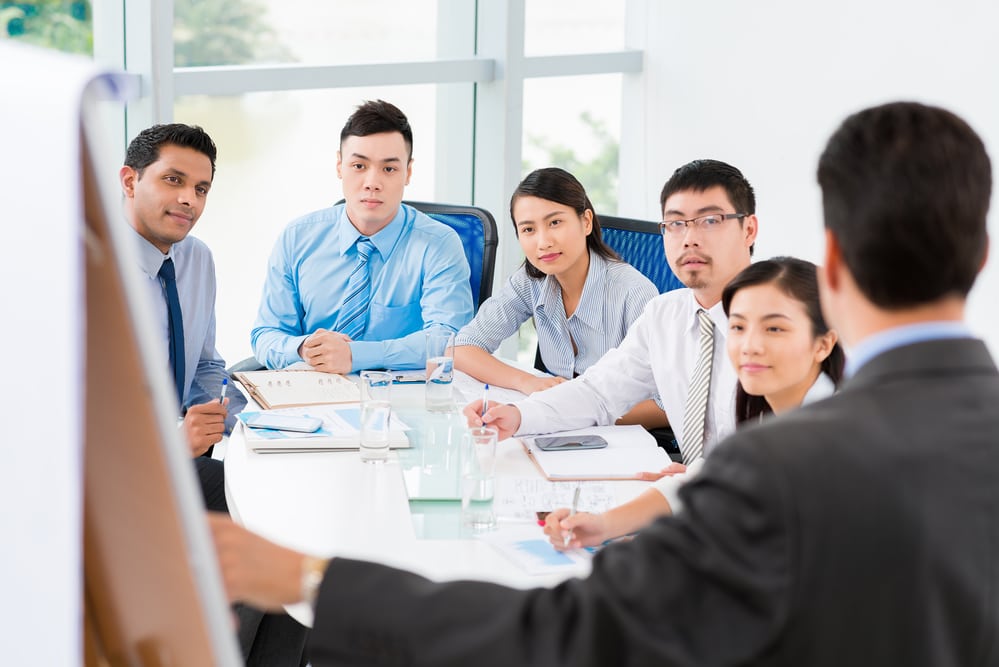 a group of work professionals sitting around an office table while a member of the staff presents a poster