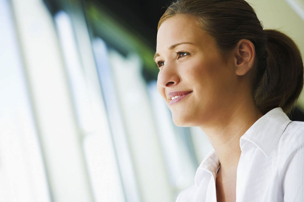 Young woman in white collared shirt smiling and looking out office window after a life change